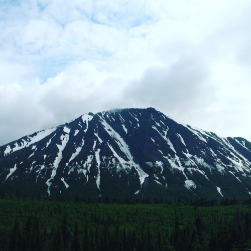 a snow-covered mountain beneath a cloudy sky and above hills covered in greenery and pine trees
