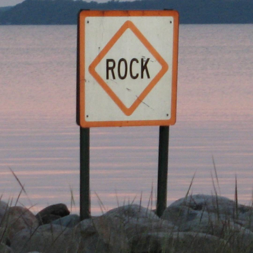 A warning sign on top of a rock near a lake. The sign is labeled with the word 'rock'